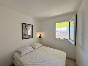 Ein Schlafzimmer mit einem Bett, einem gerahmten Bild, einer Nachttischlampe und einem Fenster mit Blick auf Gebäude.