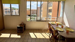 Una habitación con una mesa de comedor para cuatro personas, ventanas amplias, un pequeño estante de madera y vistas a edificios.