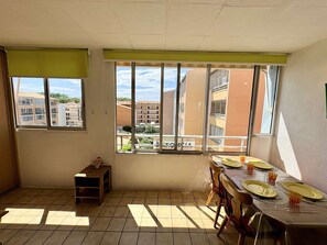 Ein Zimmer mit Esstisch, gedeckten Plätzen für vier Personen, großen Fenstern, einer kleinen Holzablage und Blick auf Gebäude.