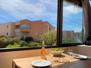 A dining table with plates, a wine glass, and a bottle of water, set against a window overlooking a residential building complex. - Loft studio, AC, beach - 2 guests (La Londe-les-Maures)