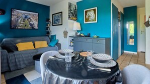 Een moderne woonkamer met een eettafel gedekt voor twee personen, een grijze bank met gele kussens, en een stadsgezicht schilderij aan de muur.