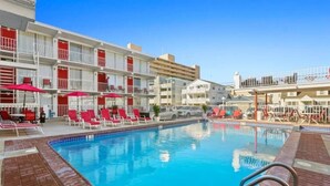 A swimming pool with red lounge chairs and a white building with balconies. - Beach Block North Wildwood | Malibu Sun @ The Malibu (North Wildwood)