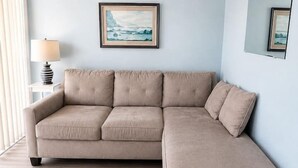 A room with a beige sectional sofa, a framed landscape painting, a side table with a lamp, and a mirror on the wall. - Beach Block North Wildwood | Malibu Sun @ The Malibu (North Wildwood)