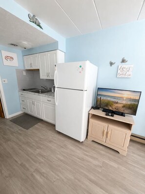 A compact kitchen with white cabinets, a white refrigerator, a small wooden cabinet with a TV, and a beach scene painting on the wall. - Beach Block North Wildwood | Malibu Sun @ The Malibu (North Wildwood)