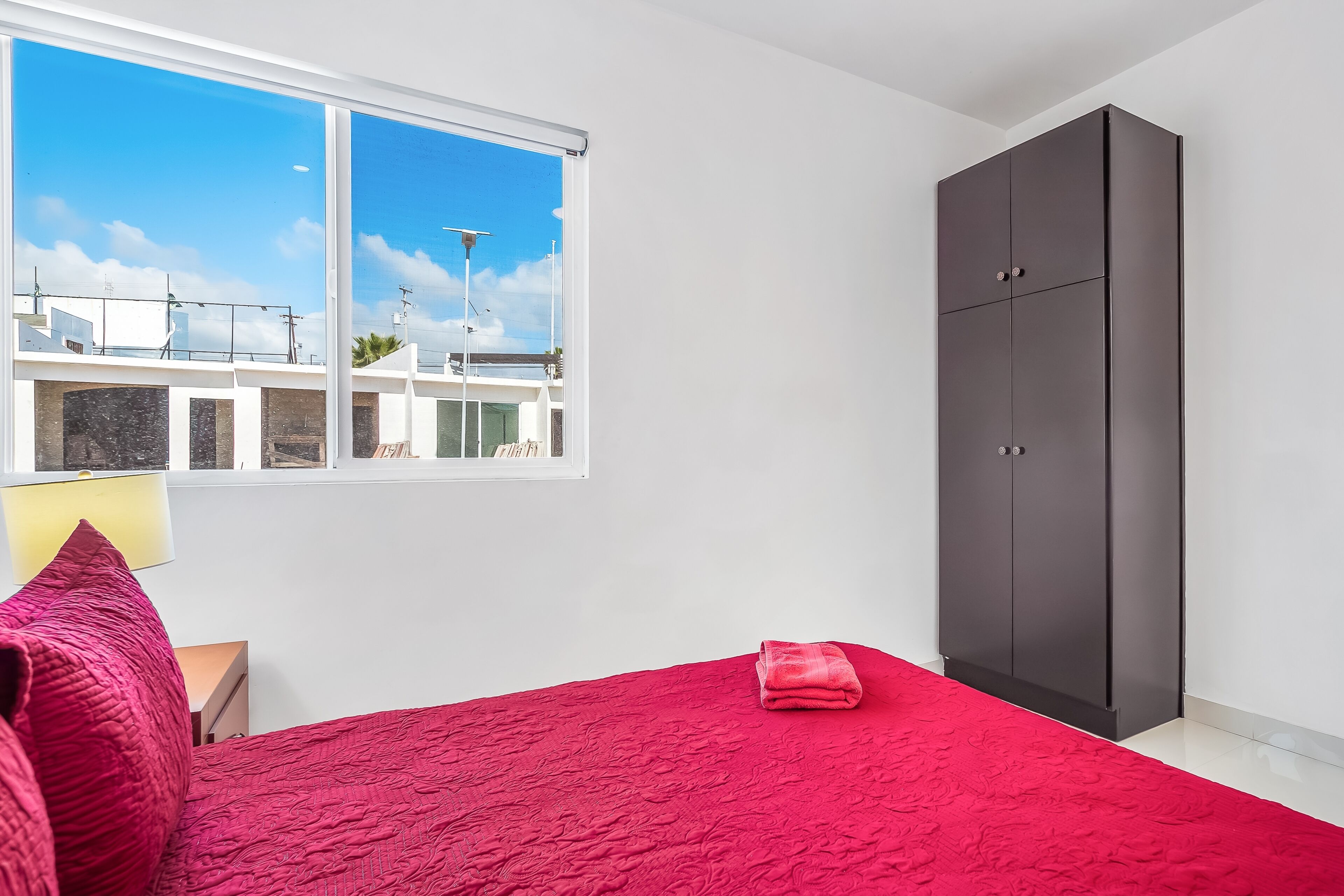 A bedroom with a red bedspread, a grey wardrobe, and a window with a view of buildings and a clear sky.