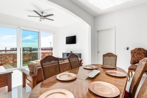 Un coin repas avec une grande table dressée pour quatre personnes, un téléviseur fixé au mur et une vue sur l’océan grâce à de grandes fenêtres.