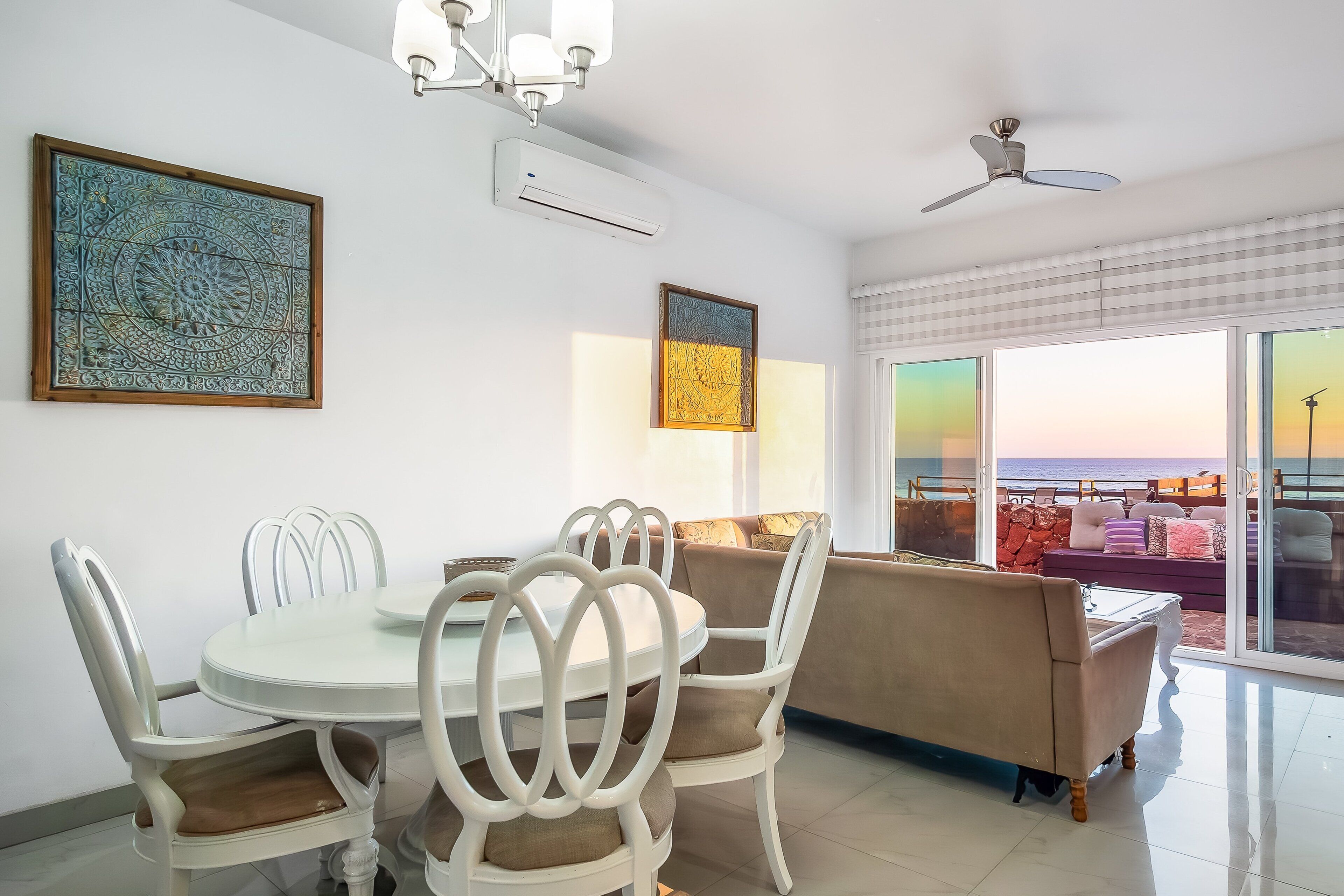 A dining area with a round table and chairs, a ceiling fan, and a large window with a view of the ocean.