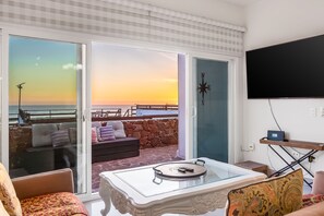 A room with a glass table, a sofa, a TV, and a view of the sunset over the sea.