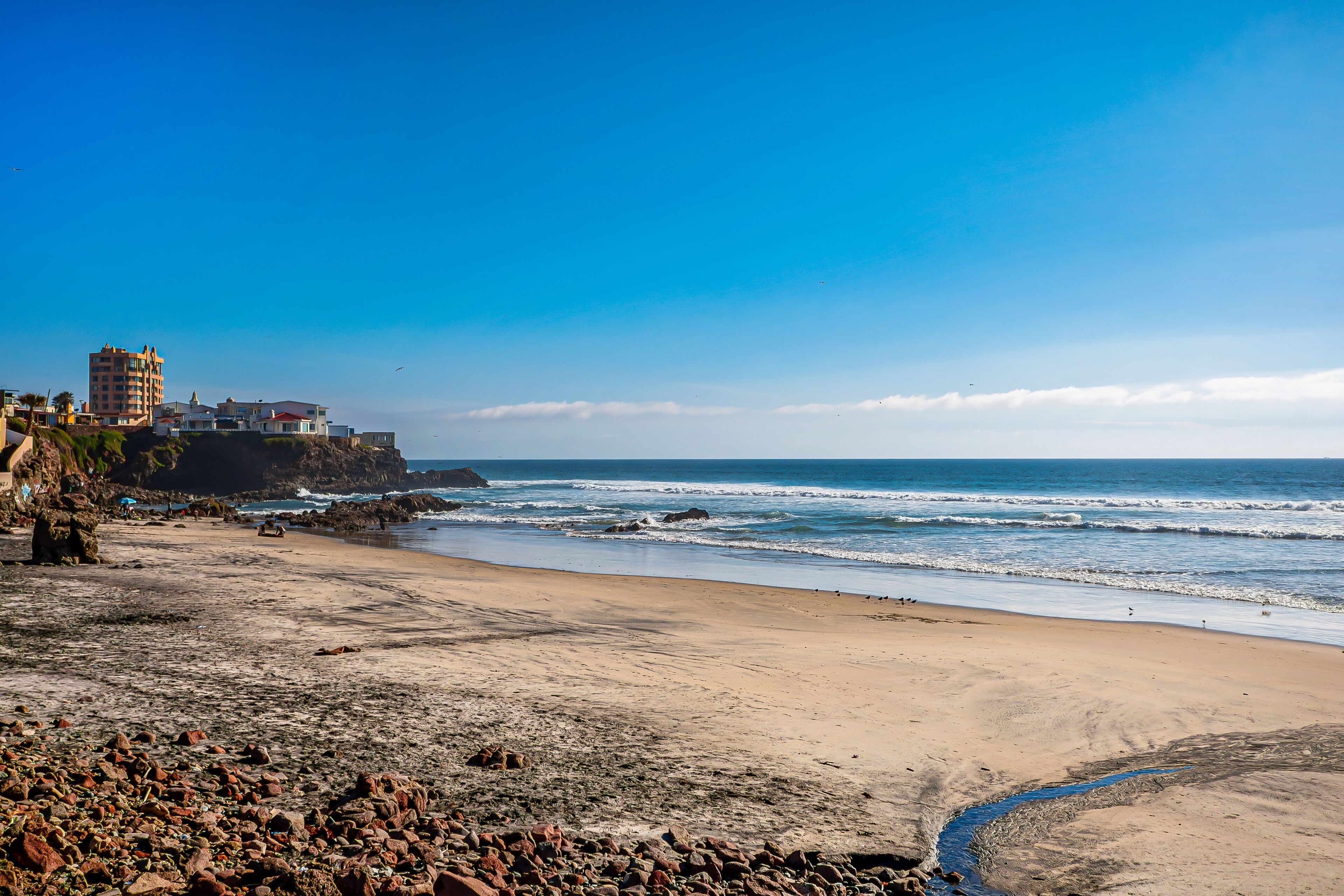 Uma praia com um primeiro plano rochoso, um edifício no alto de um penhasco e o oceano ao fundo.