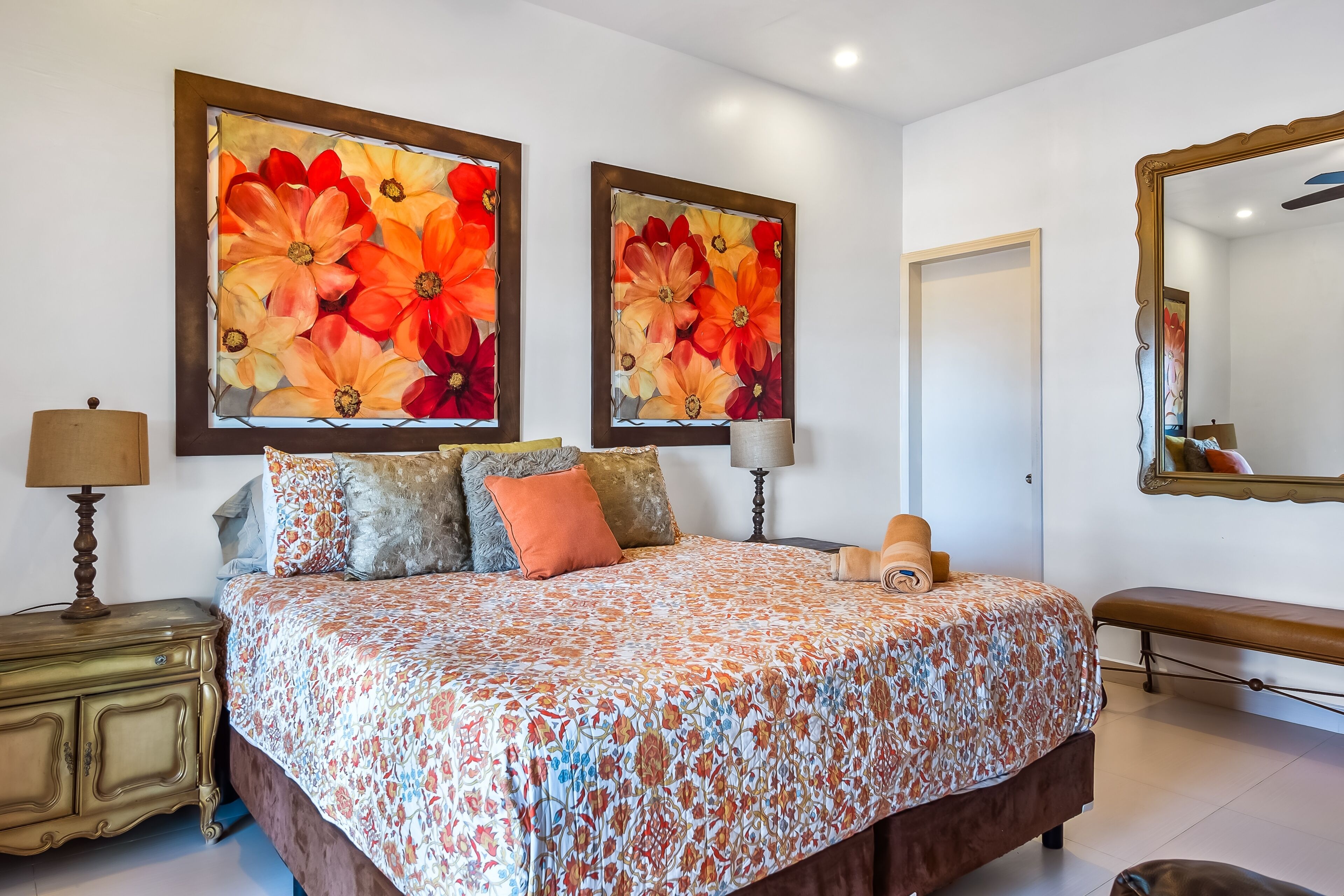Un dormitorio con una cama grande, dos cuadros de flores en la pared, una lámpara de noche, un banco y un espejo.