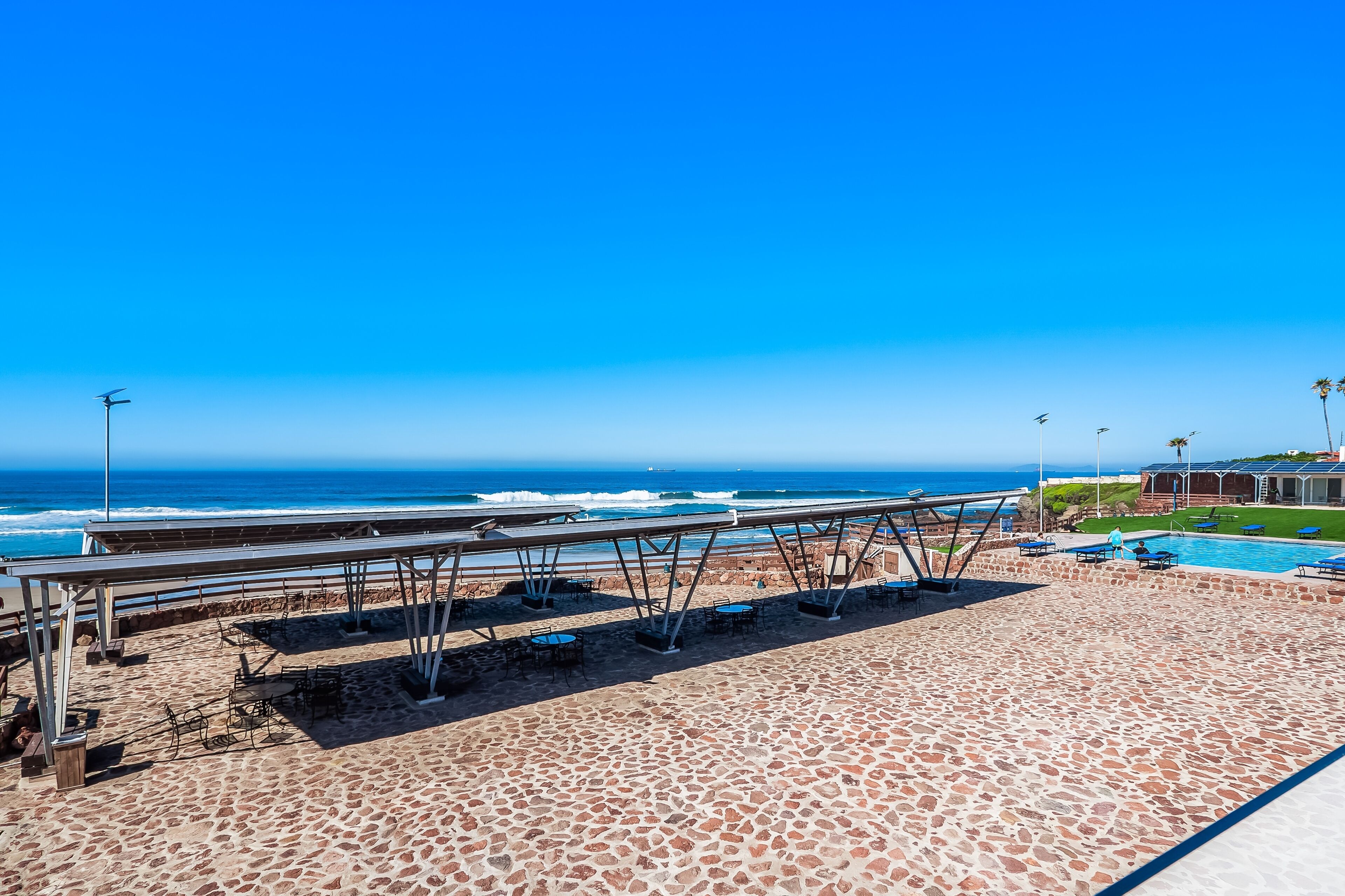 A beachside patio with a view of the ocean, a pool, and a building in the background.