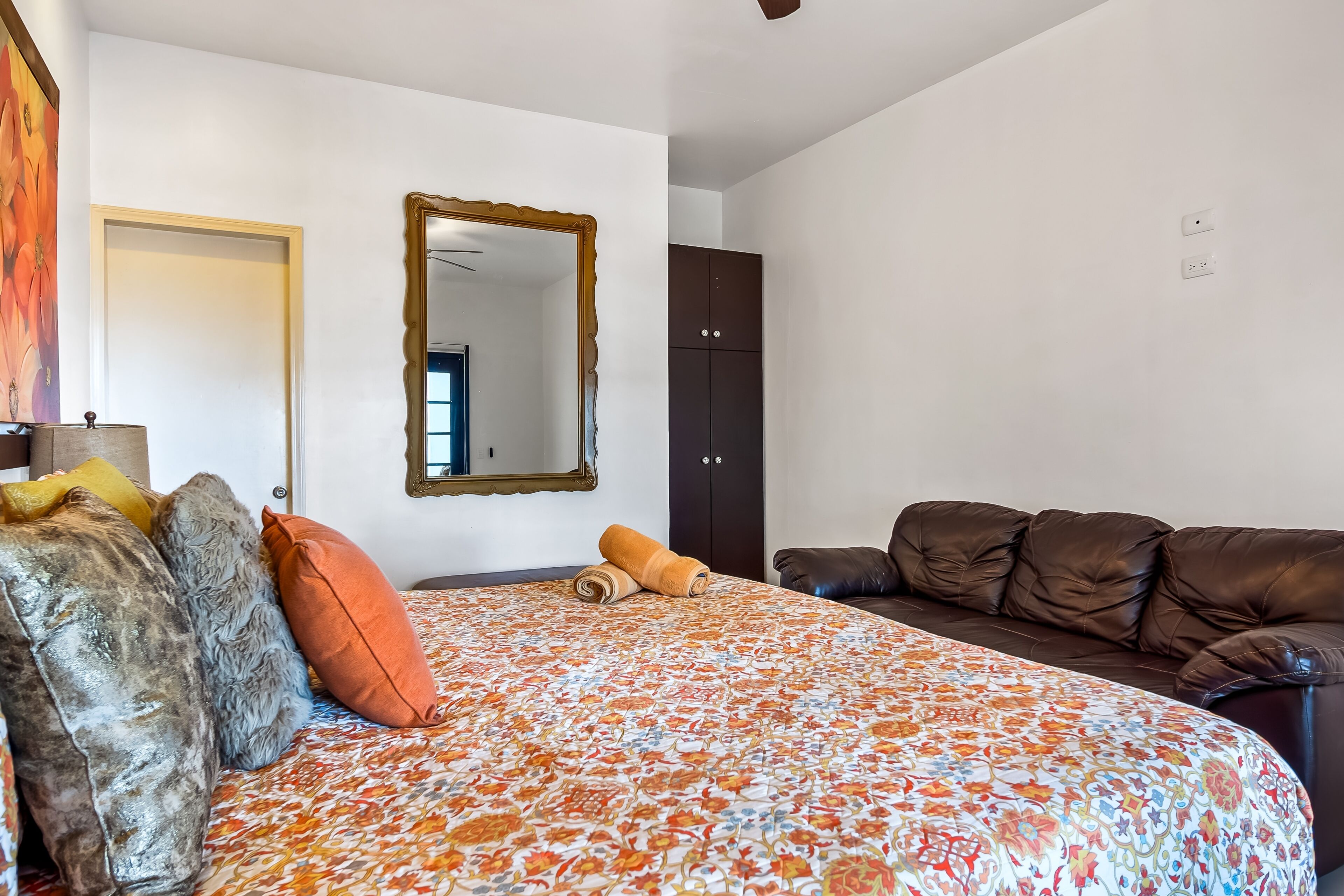Un dormitorio con una cama, un sofá de cuero marrón, un armario oscuro y un espejo en la pared.