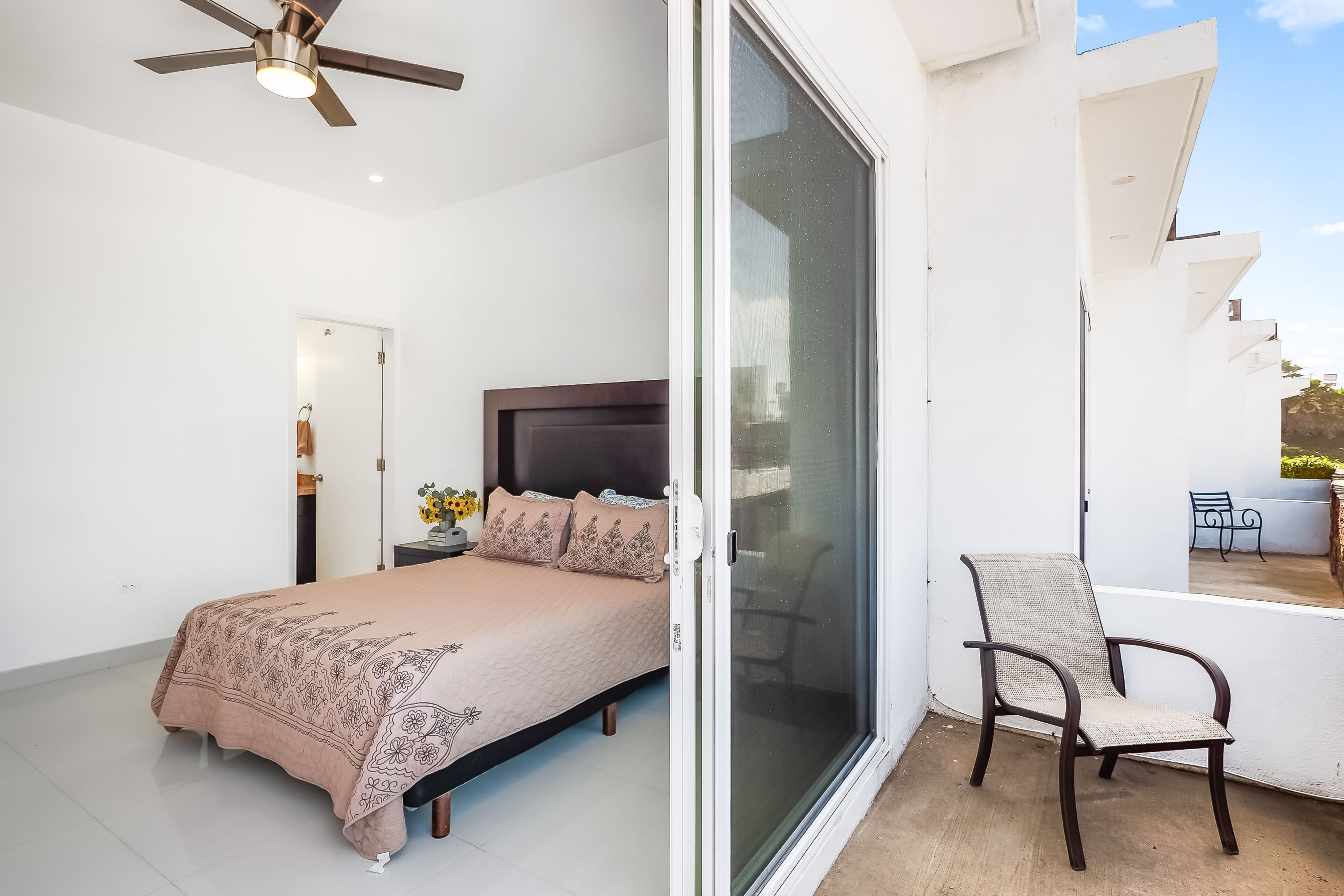 Une chambre à coucher comprenant un lit, un ventilateur de plafond et une porte-fenêtre coulissante donnant sur un espace extérieur avec un fauteuil.