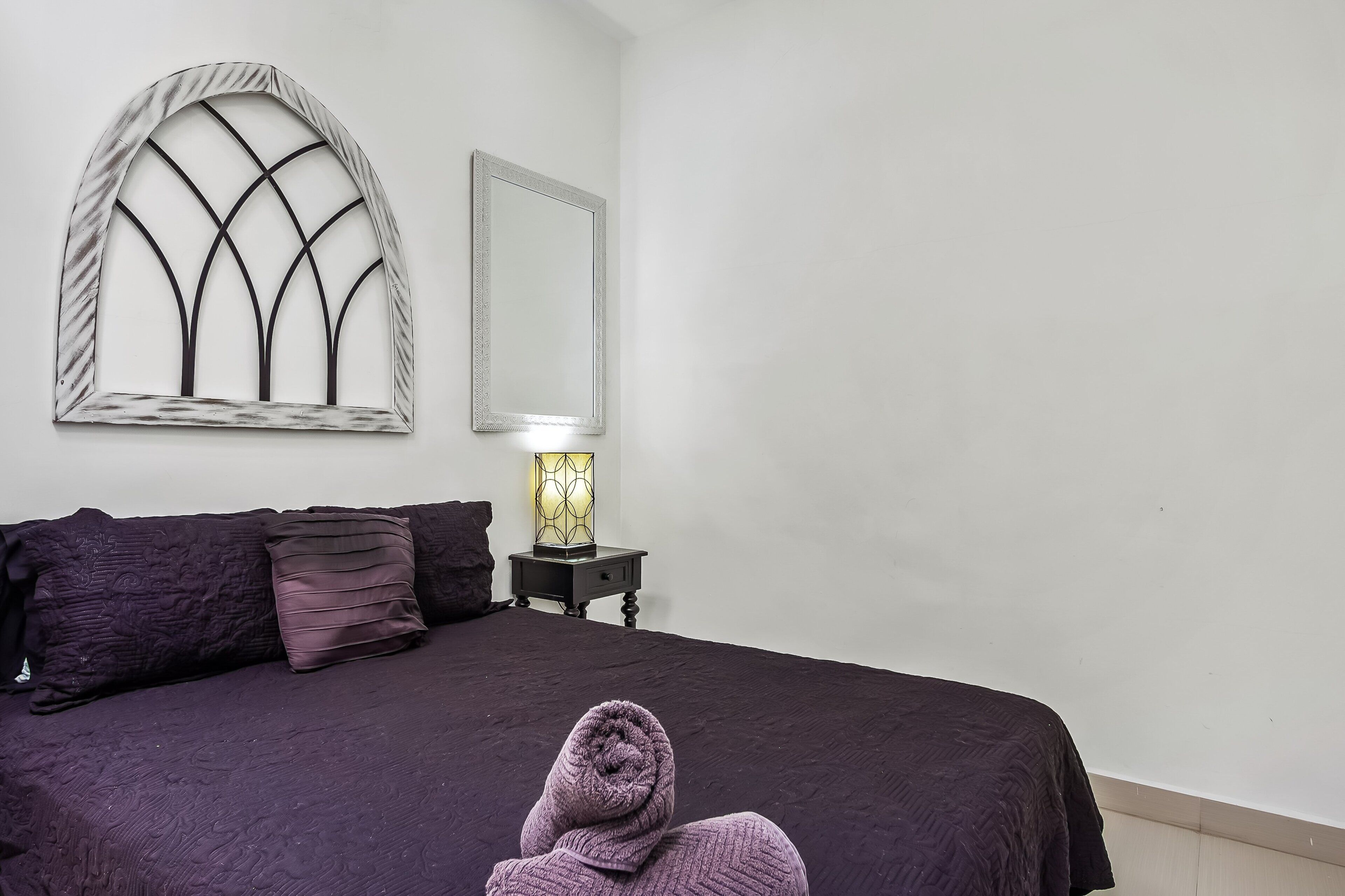 Un dormitorio con una cama, una mesita de noche con lámpara y un ventanal decorativo.
