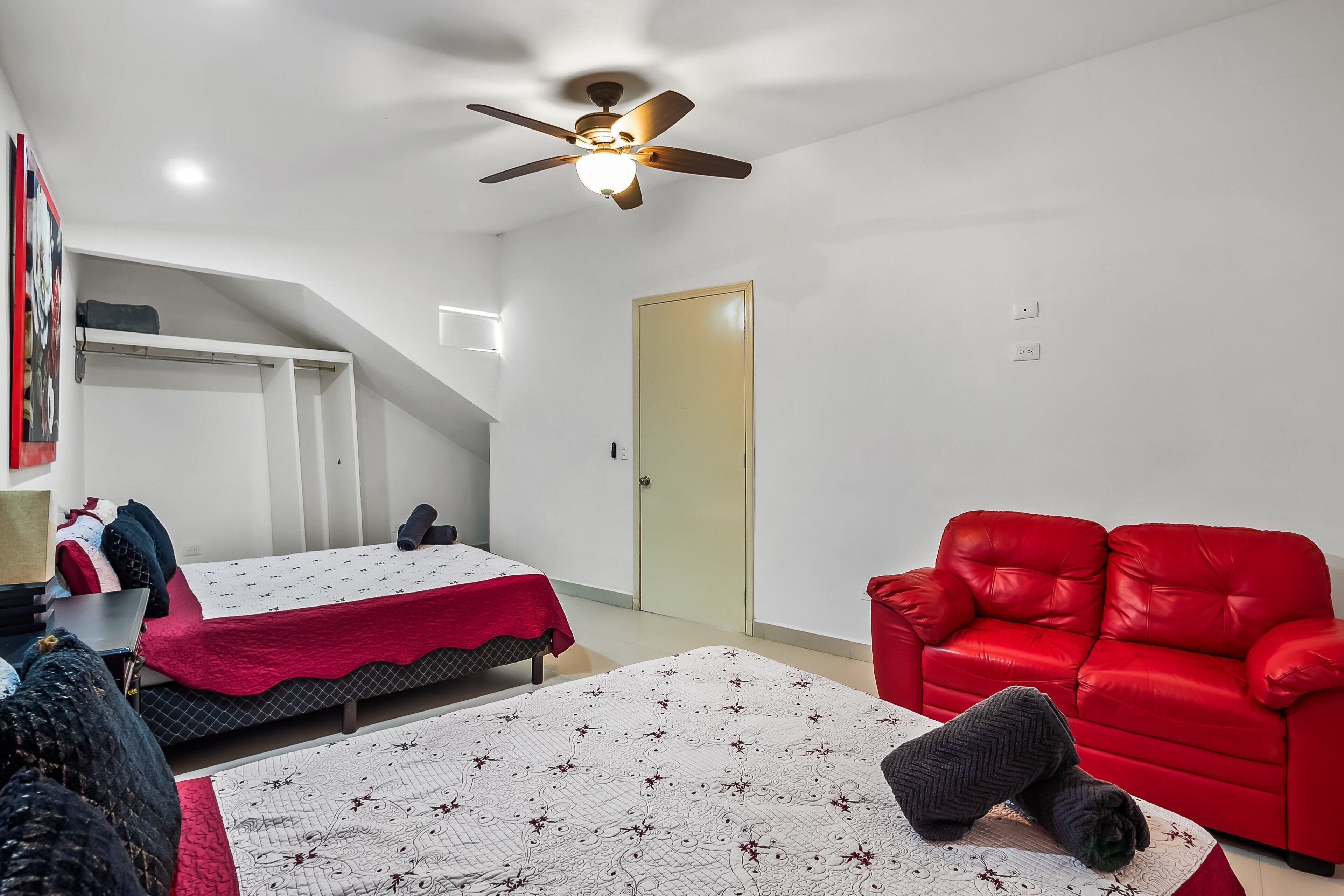 A bedroom with a bed, a red sofa, a ceiling fan, and a door.