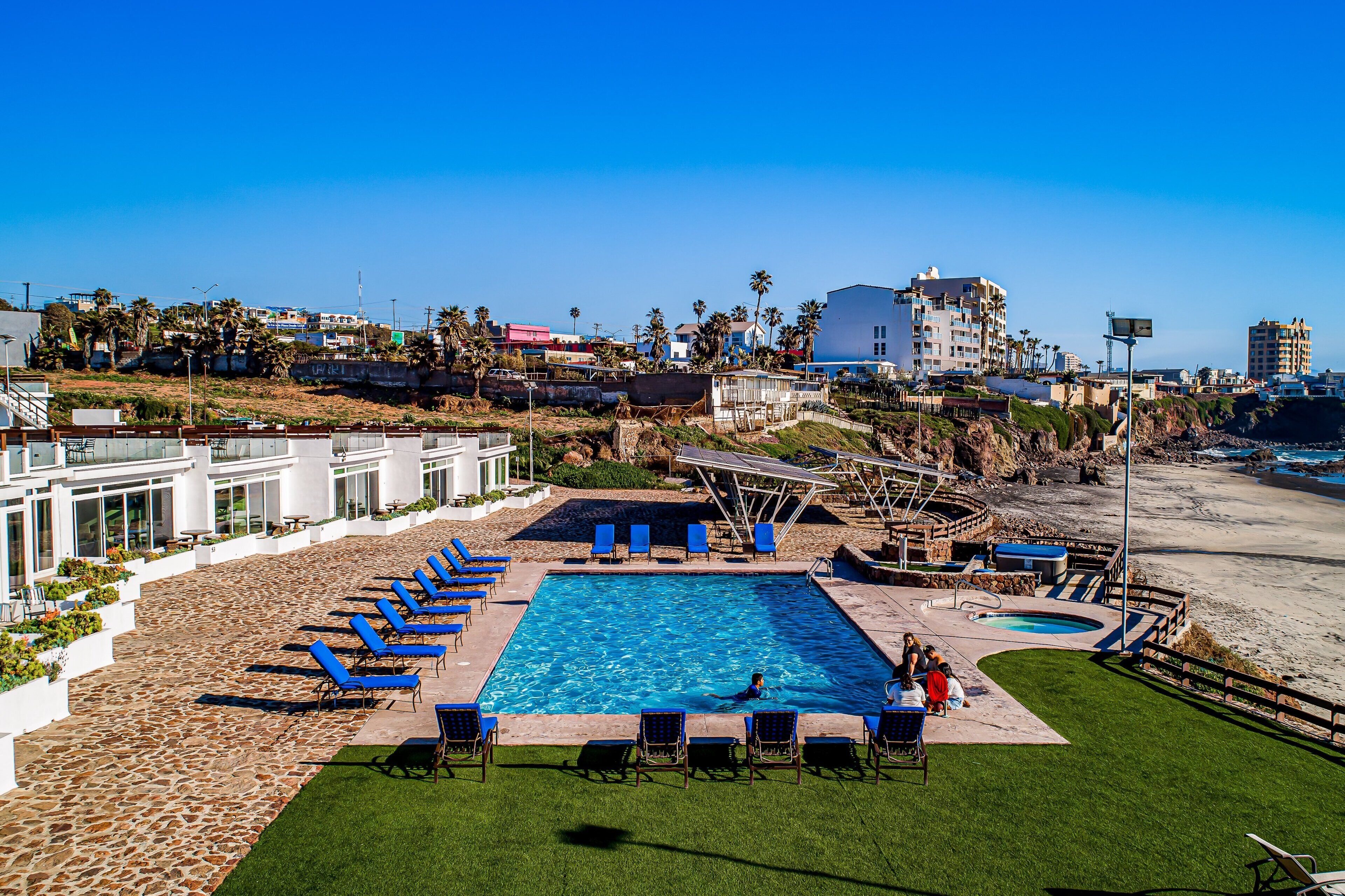 A swimming pool with lounge chairs, a small hot tub, and a view of the beach and buildings in the background.