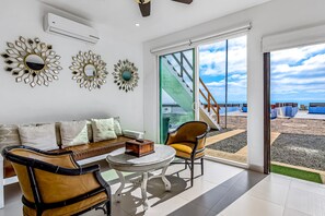 Ein modernes Wohnzimmer mit einer Couch, Sesseln, einem Tisch, dekorativen Spiegeln und einer großen Schiebeglastür, die auf einen Balkon mit Meerblick führt.