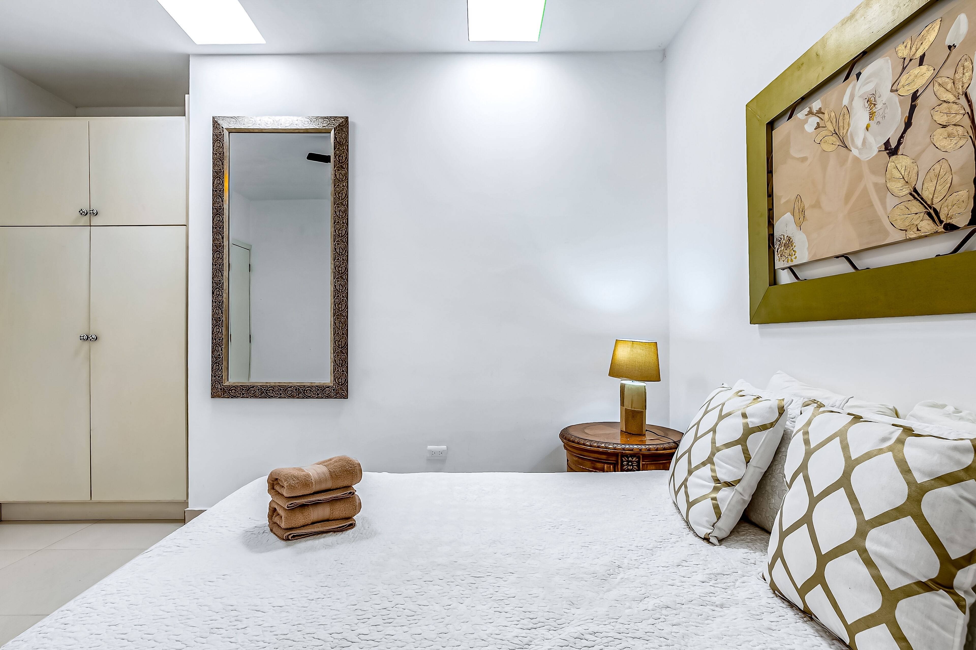 Een slaapkamer met een bed, een kledingkast, een spiegel, een lamp en een kunstwerk in een lijst aan de muur.