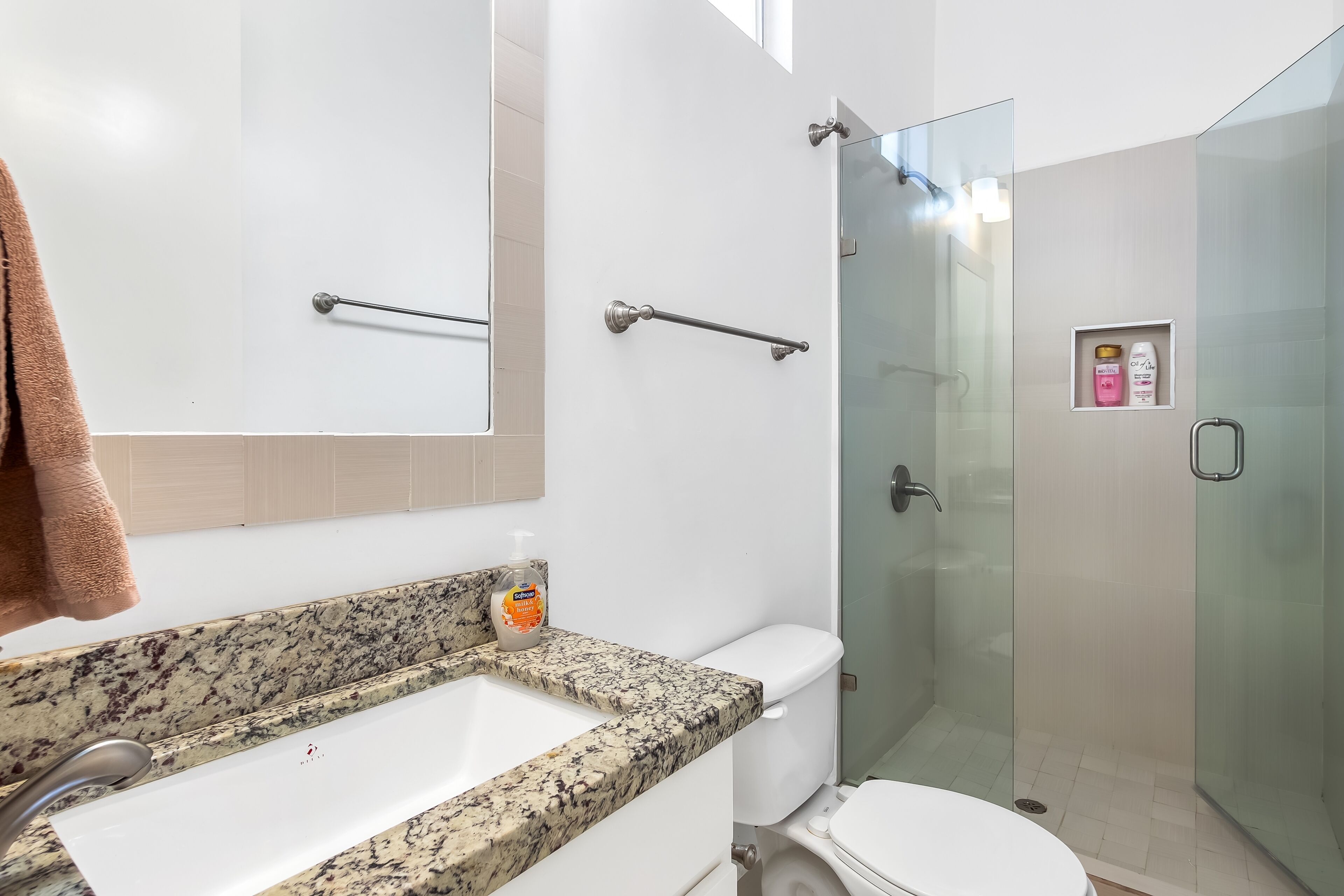 Une salle de bain dotée d’un comptoir en granit, d’un lavabo blanc, de toilettes et d’une douche à parois de verre.