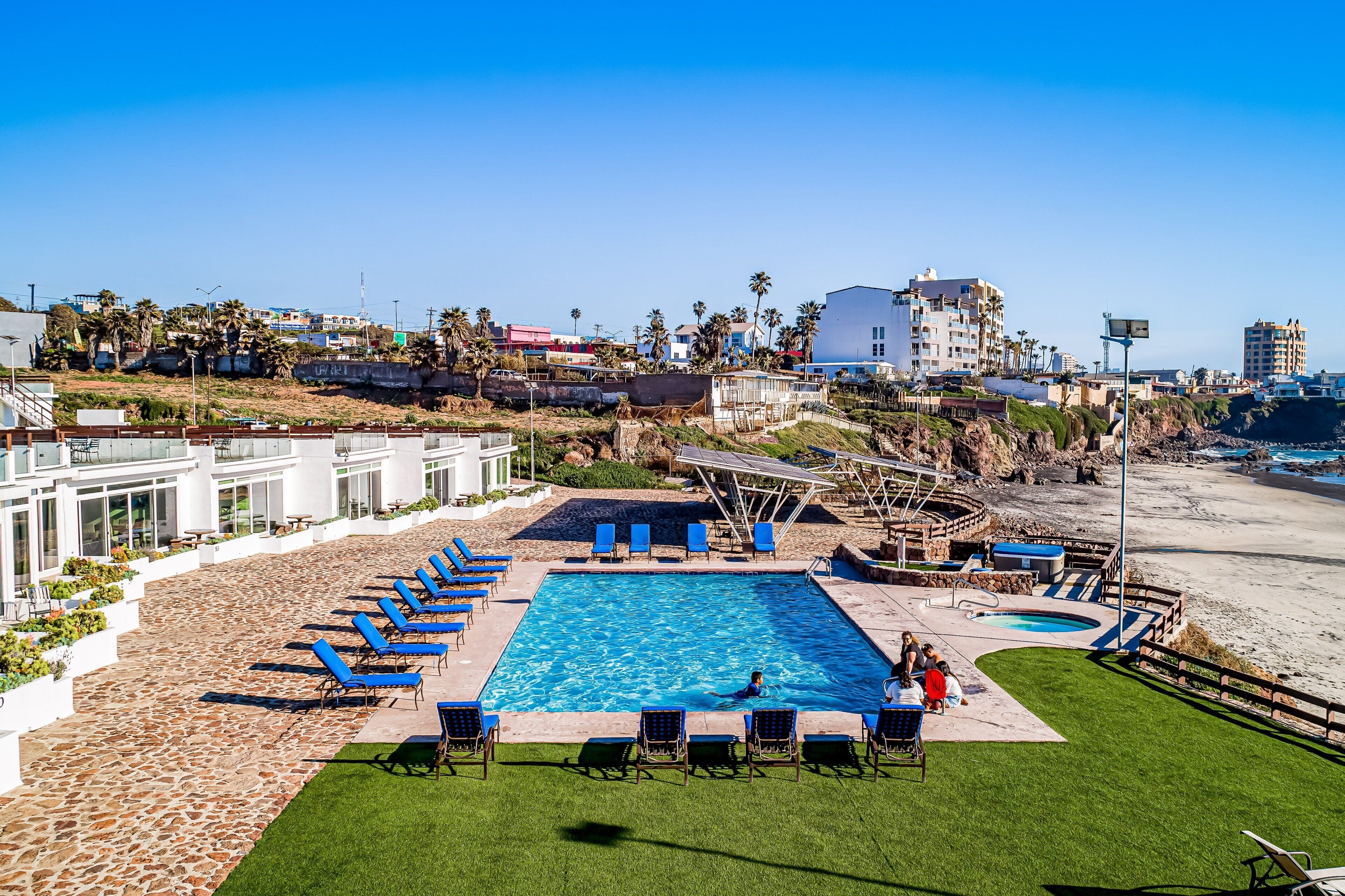 A swimming pool with lounge chairs, a small hot tub, and a view of the beach and buildings.