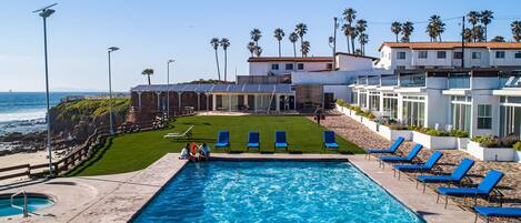 A swimming pool with lounge chairs, a grassy area, and a building with a view of the ocean.