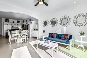 A modern living room with a white sofa, a dining table, and a kitchen in the background. - Dog-friendly Oceanfront Villa With Rooftop Patio, Pool Access, & Kitchen (Playas de Rosarito)