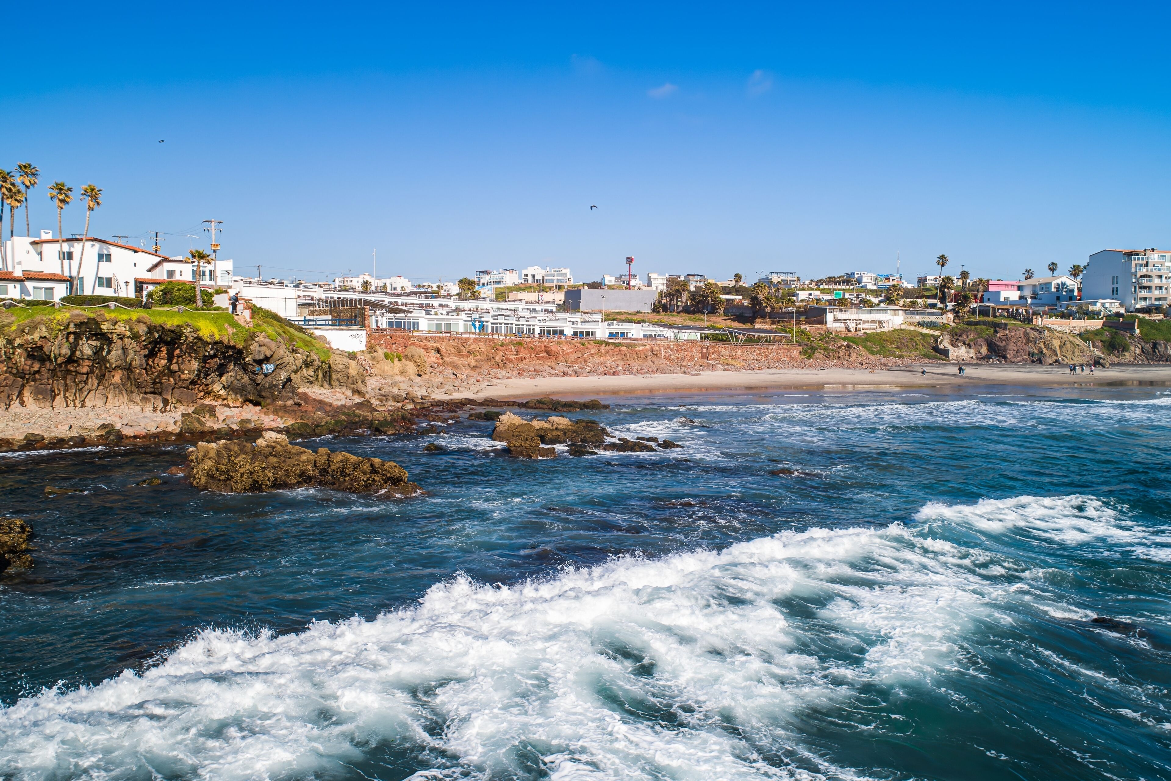 A coastal view with rocky shoreline, waves, and buildings in the background.