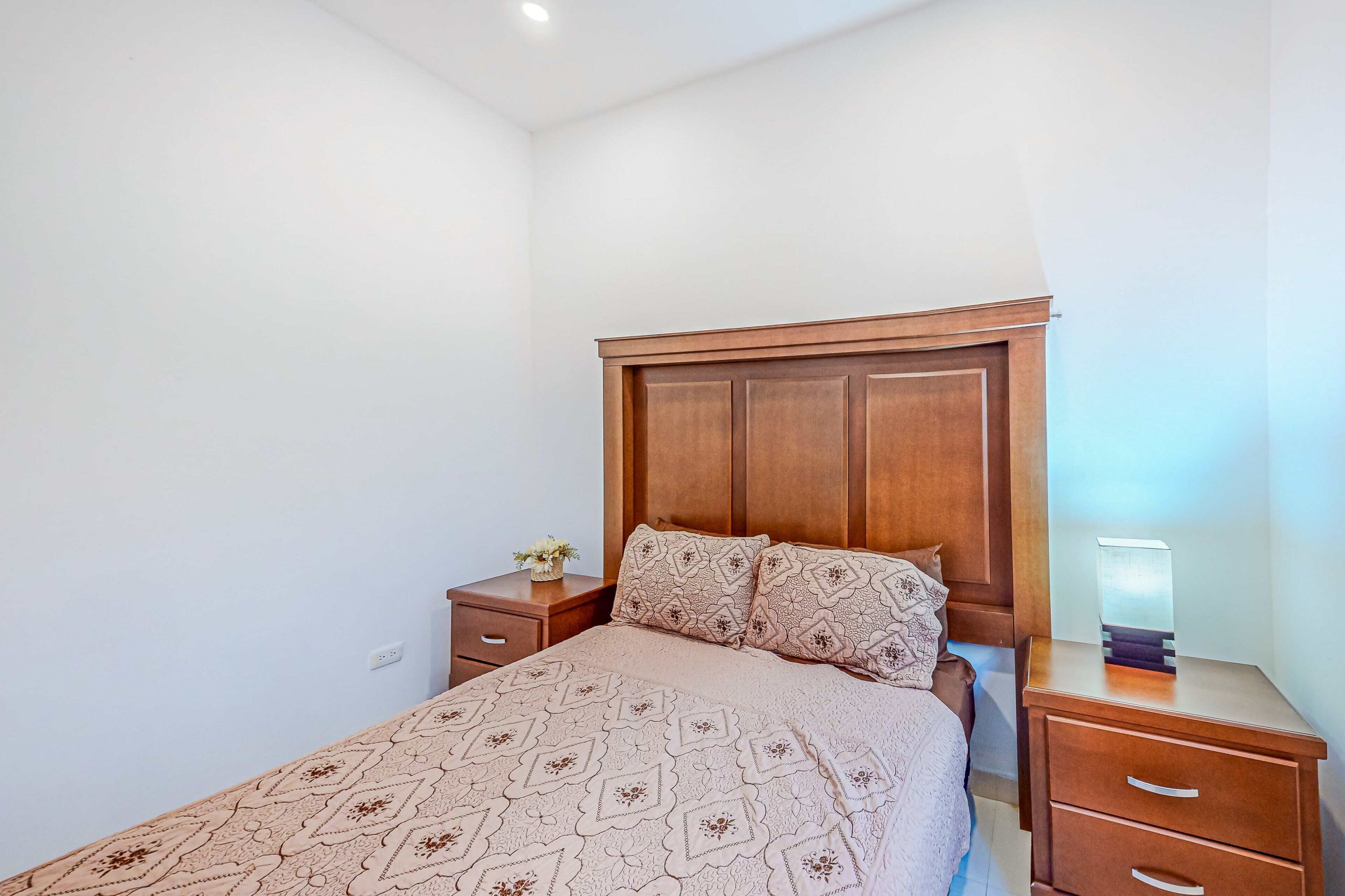 Un dormitorio con cabecera de madera, colcha estampada, mesita de noche con lámpara y una mesita auxiliar con un florero.