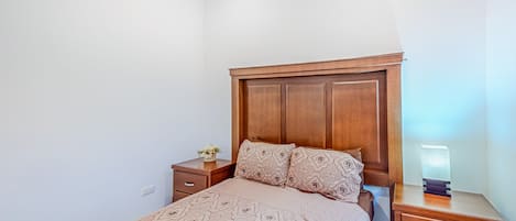 Un dormitorio con cabecera de madera, colcha estampada, mesita de noche con lámpara y una mesita auxiliar con un florero.