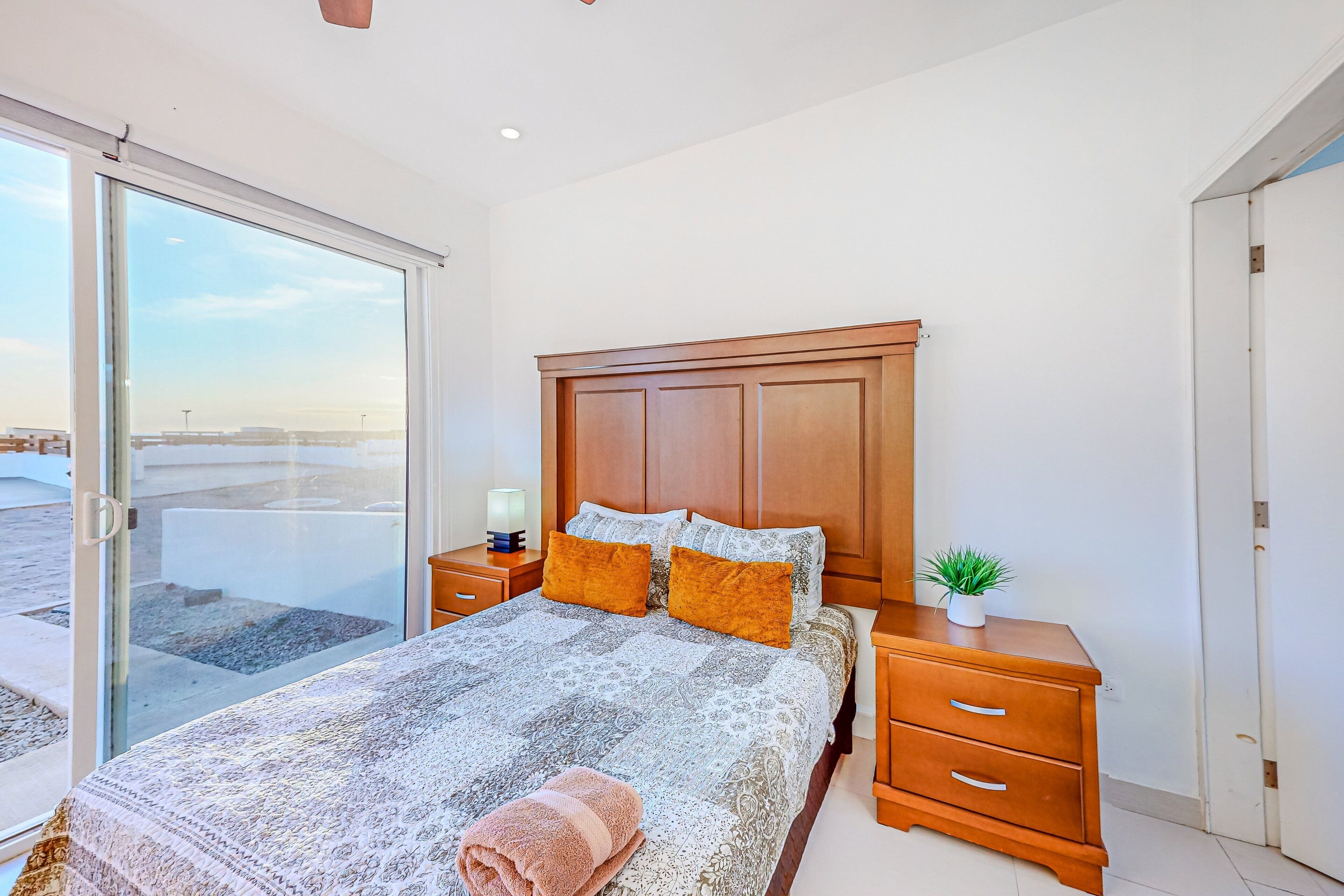 A bedroom with a large bed, wooden headboard, two bedside tables, a small plant, and a sliding glass door leading to an outdoor area.