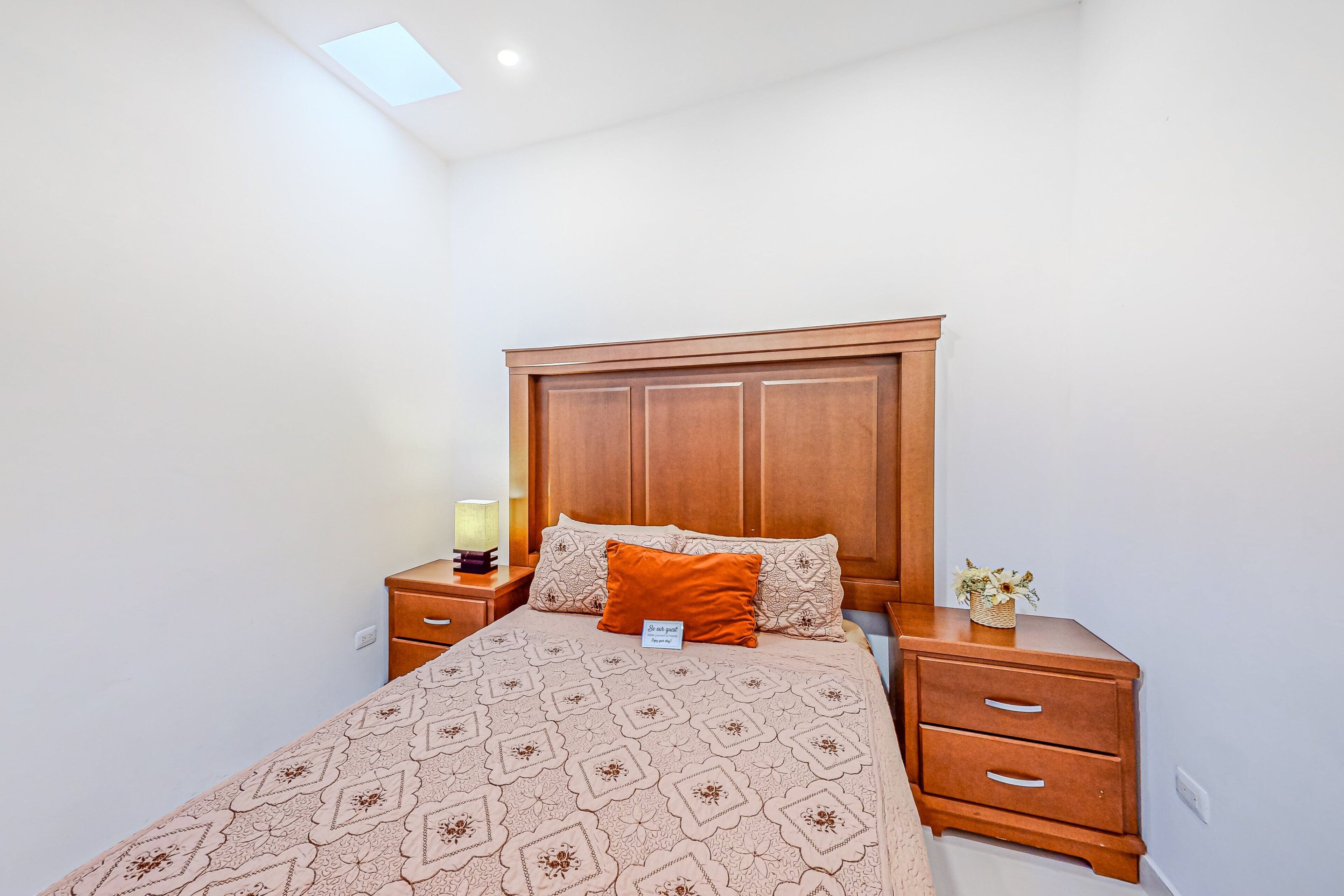 A bedroom with a wooden headboard, a patterned bedspread, a single orange pillow, a bedside table with a lamp, and a small plant.