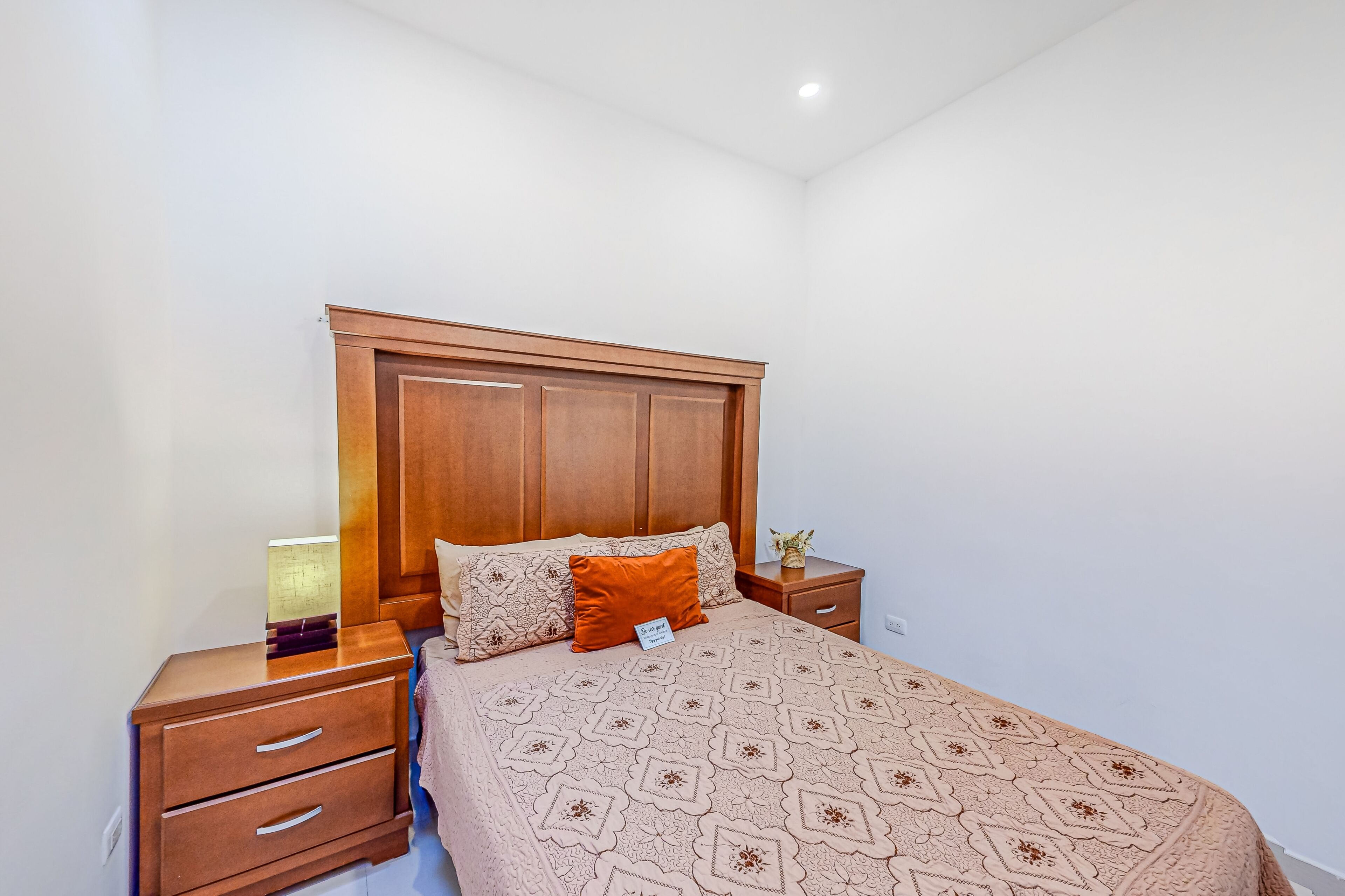 Un lit bien fait, recouvert d’une couvre-lit à motifs, avec une tête de lit en bois et une table de chevet dotée de tiroirs et d’une lampe.