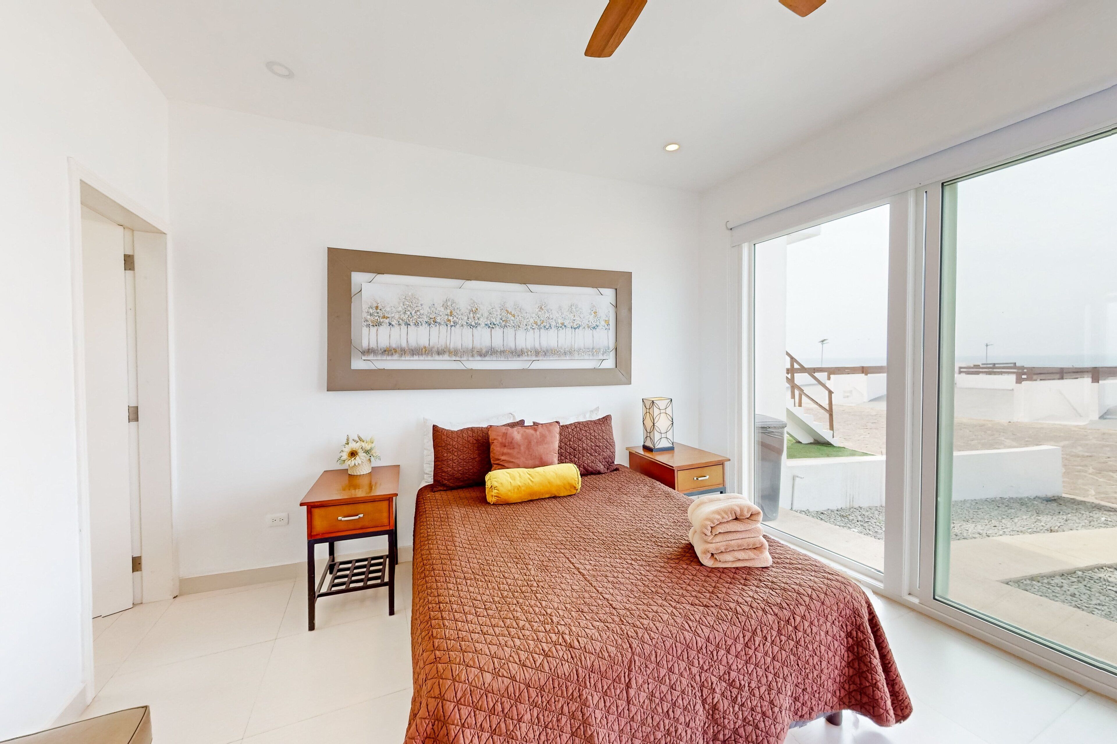 Quarto com cama grande, mesa de cabeceira, obra de arte emoldurada e vista para a praia através de uma janela ampla.