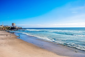 A beach with a clear sky, a few people walking, and a building with a tower on the left. - Dog-friendly Beachfront Retreat With Rooftop Deck, Pool, Hot Tub, & Kitchen (Playas de Rosarito)