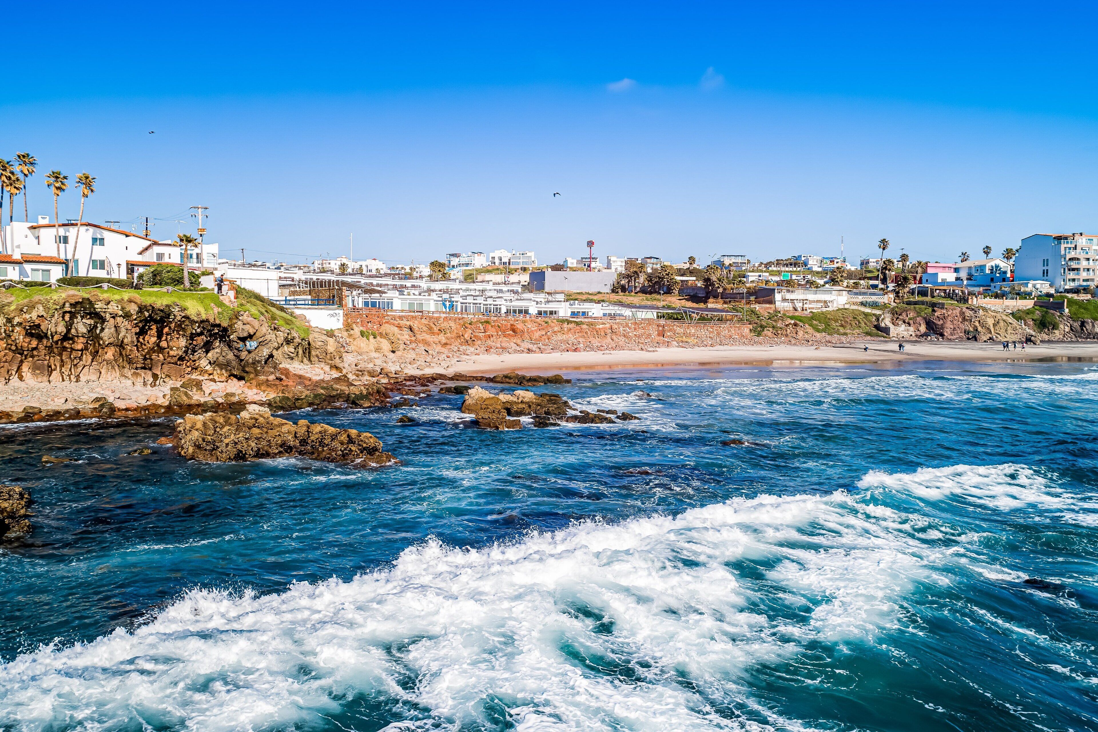A coastal view with rocky shoreline, waves, and buildings in the background.