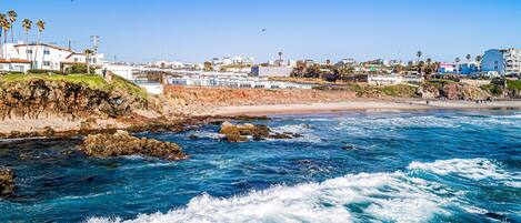 Vista costeira com linha de costa rochosa, ondas e edifícios ao fundo.