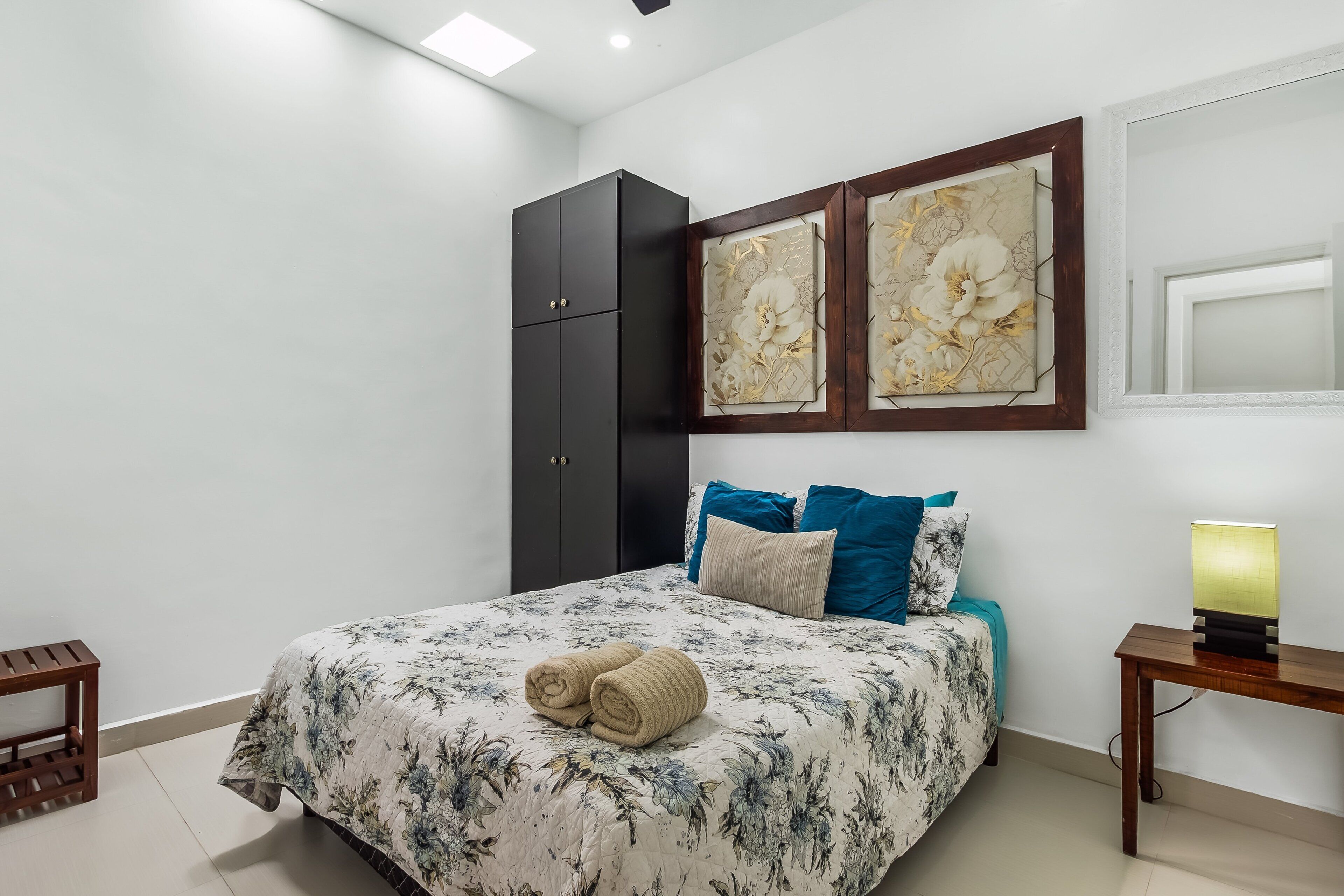 A bedroom with a bed, a wardrobe, two wall-mounted artworks, a bedside table with a lamp, and a small wooden stool.