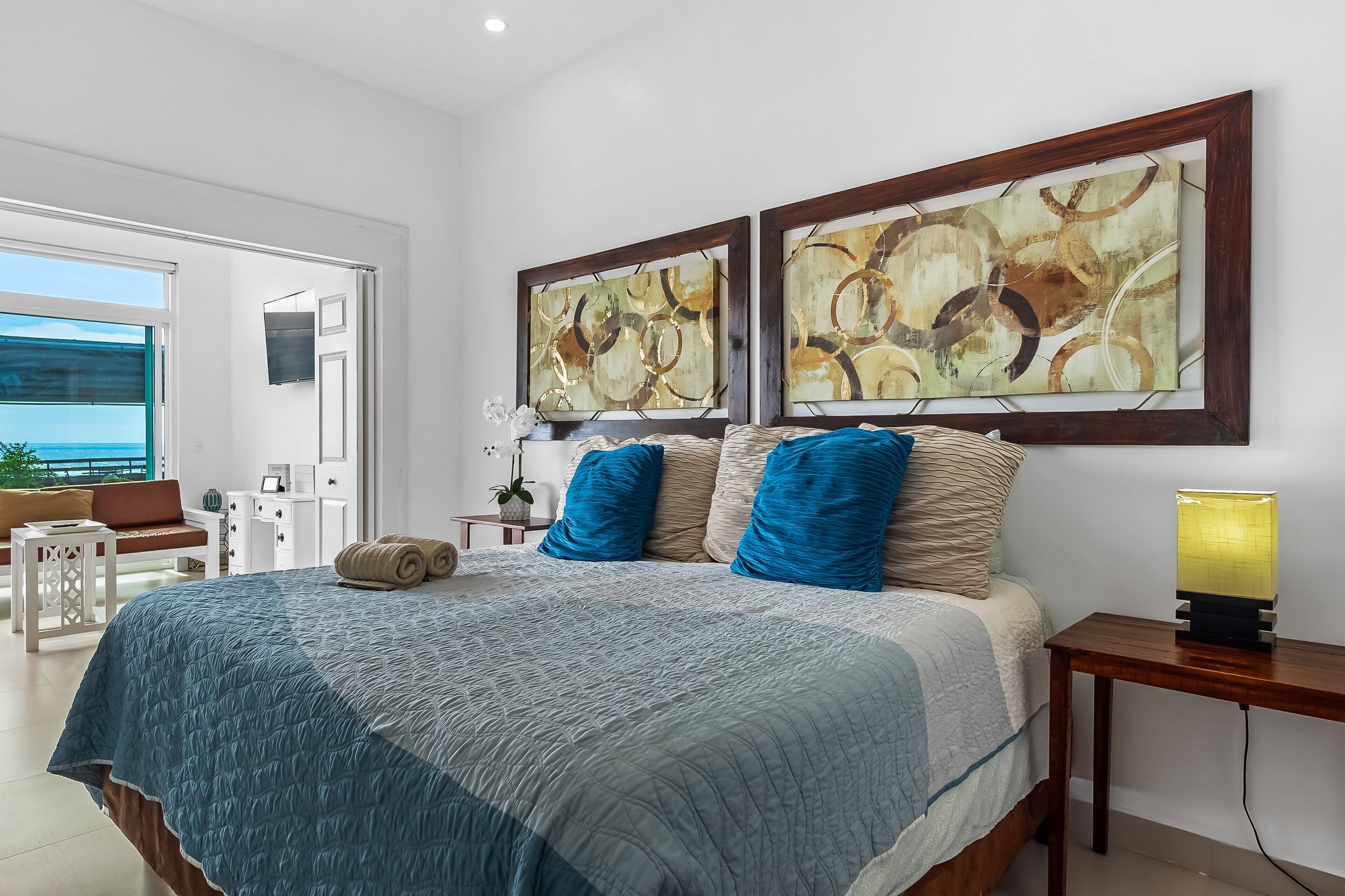 A bedroom with a bed, two wall-mounted paintings, a bedside table with a lamp, and a view of the outside.
