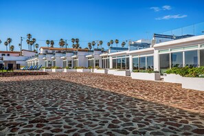 A modern hotel with a paved courtyard, palm trees, and a clear blue sky. - Dog-friendly Home With Heated Pool, Hot Tub, Partial Ocean Views, & Balcony (Playas de Rosarito)