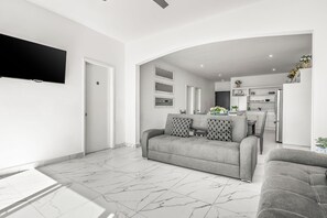 A modern living room with a grey sofa, a flat-screen TV, and a kitchen in the background. - Dog-friendly Home With Heated Pool, Hot Tub, Partial Ocean Views, & Balcony (Playas de Rosarito)