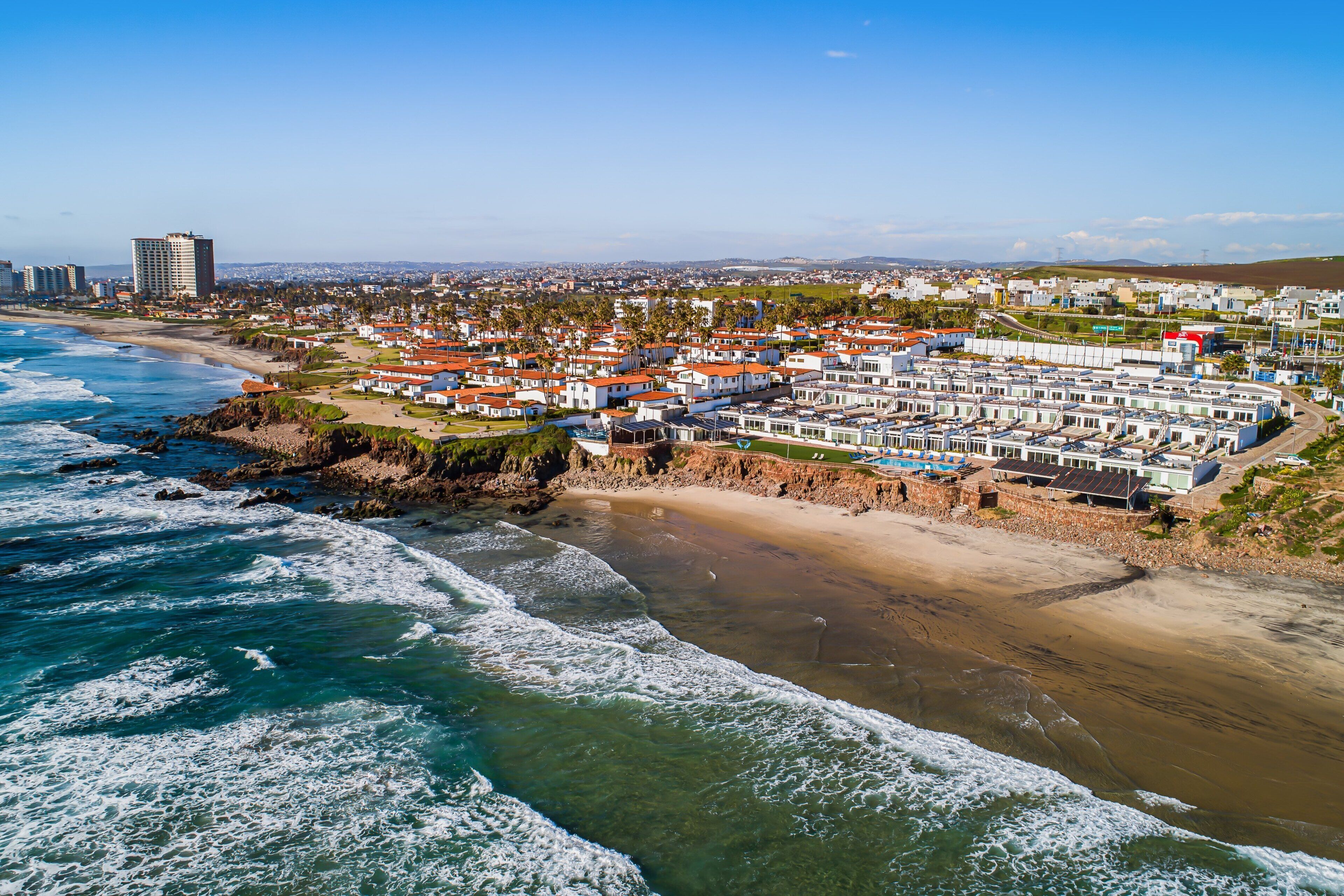 A coastal area with a beach, ocean waves, and a row of white buildings with red roofs.