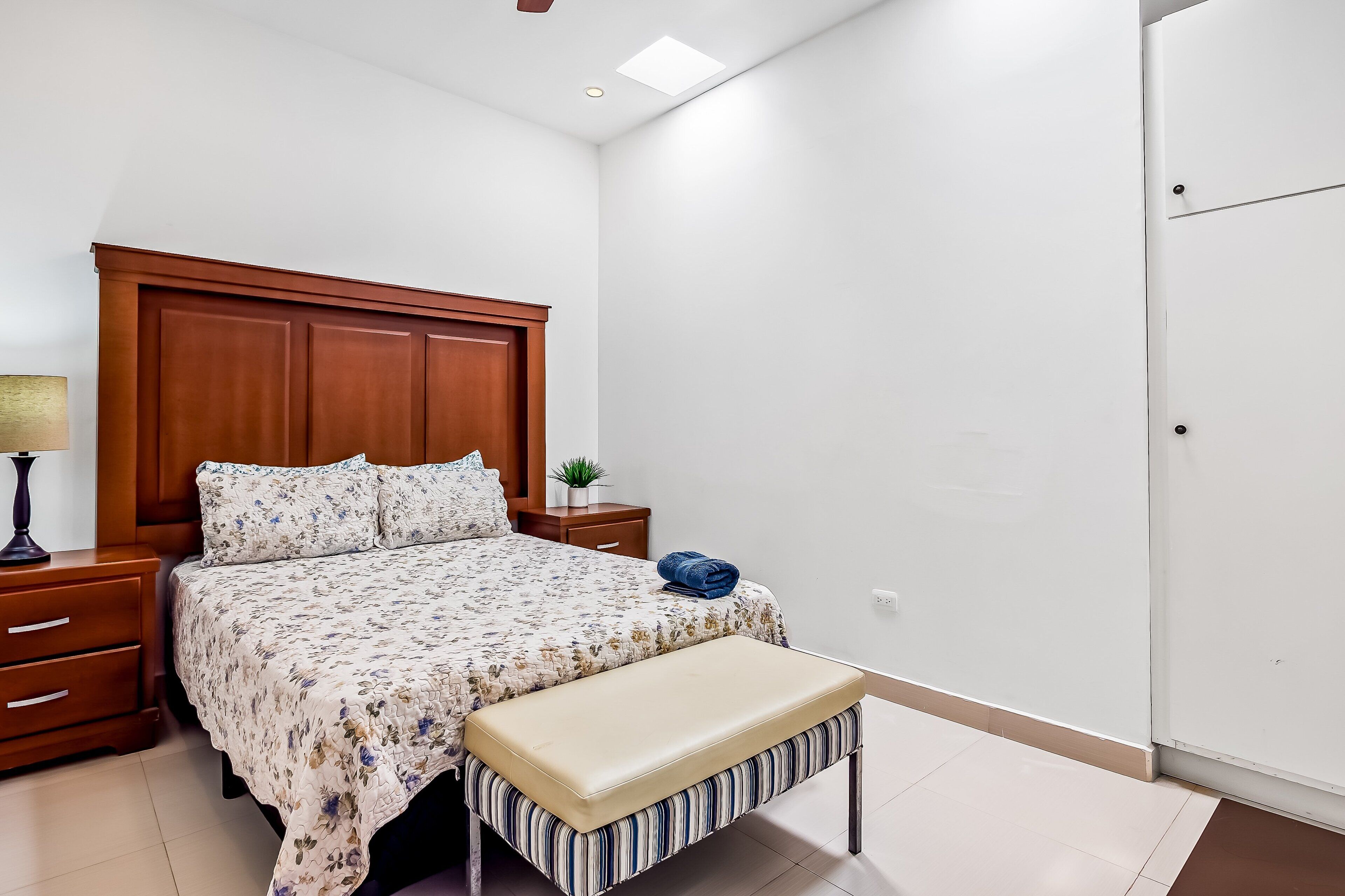 A bedroom with a wooden headboard, a bed with floral bedding, a bench, a nightstand with a lamp, and a small plant.