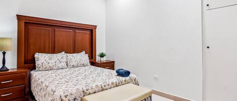 A bedroom with a wooden headboard, a bed with floral bedding, a bench, a nightstand with a lamp, and a small plant.