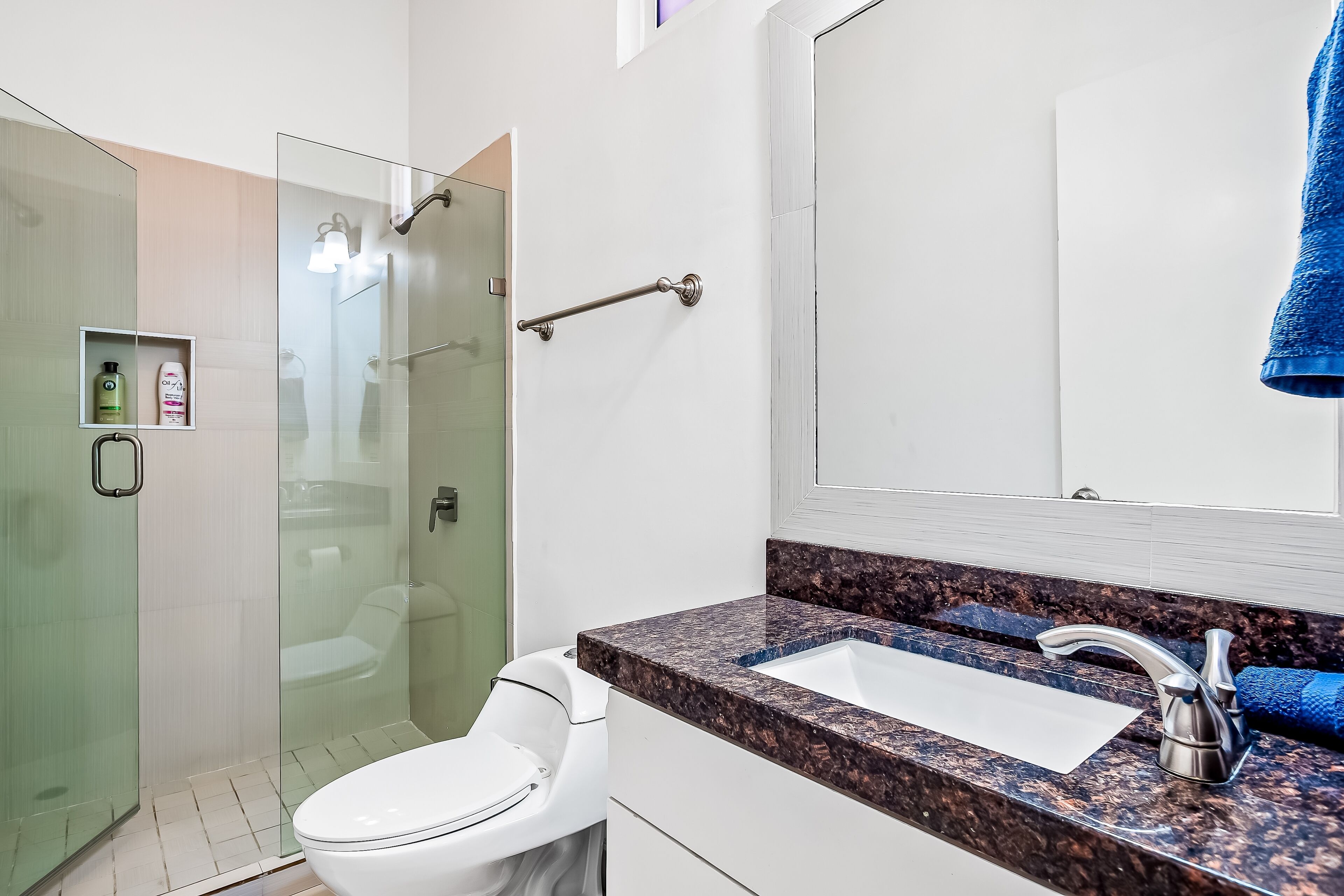A modern bathroom with a glass shower enclosure, a white toilet, a granite countertop sink, and a blue towel hanging on the wall.