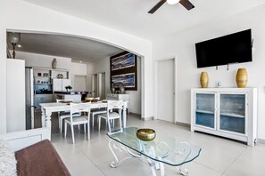 Uma sala de estar moderna com uma mesa de centro de vidro, conjunto de jantar branco e uma TV montada na parede.
