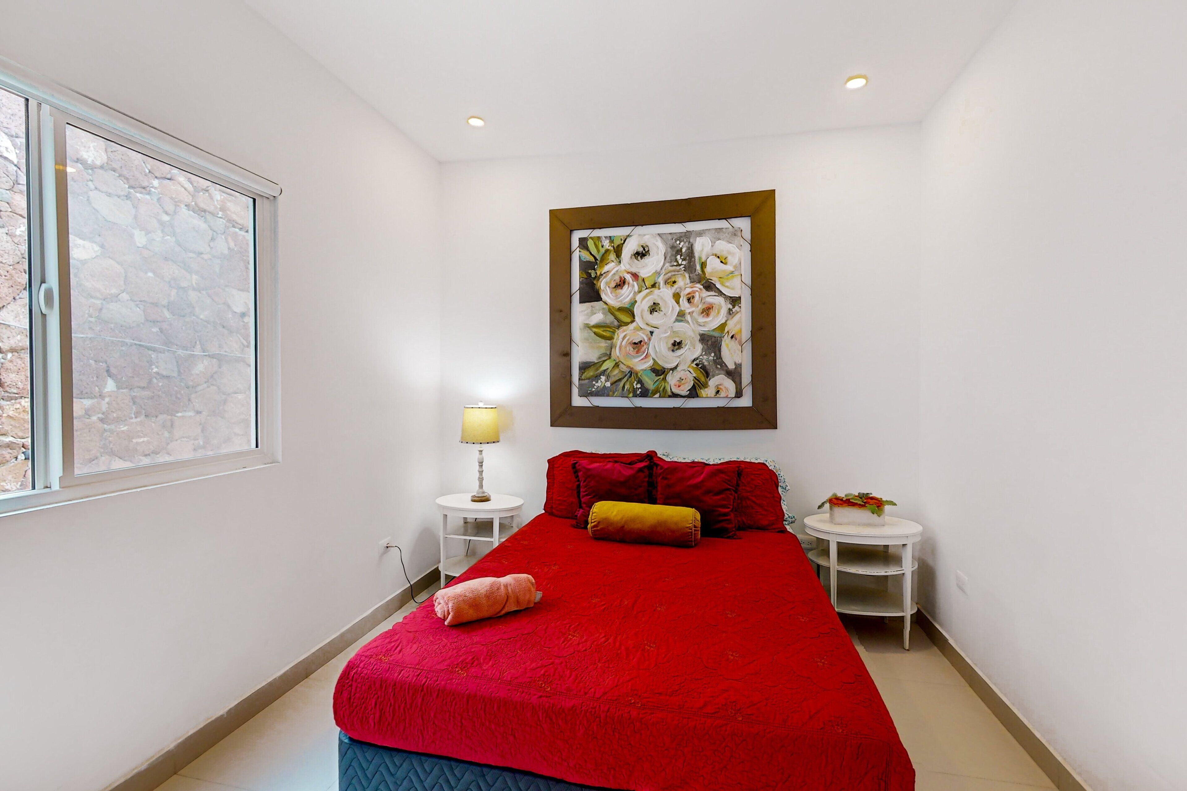 A bedroom with a red bedspread, a painting above the bed, a lamp on a bedside table, and a small table with a plant.