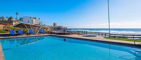 A clear blue pool with blue lounge chairs, a lamp post, and a view of the ocean and buildings in the background.