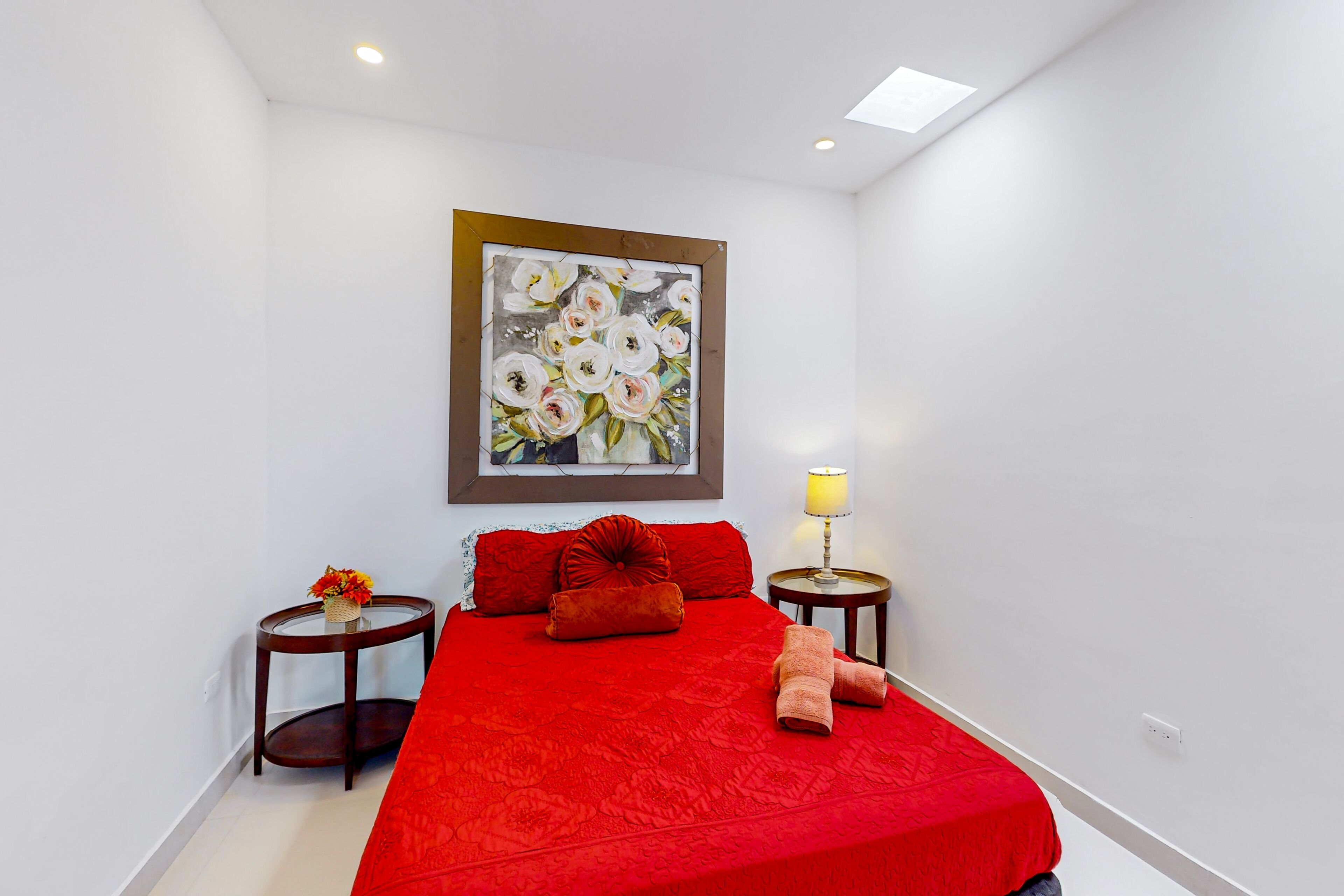 A bedroom with a red bedspread, a painting on the wall, a round table with a lamp, and a vase with flowers.