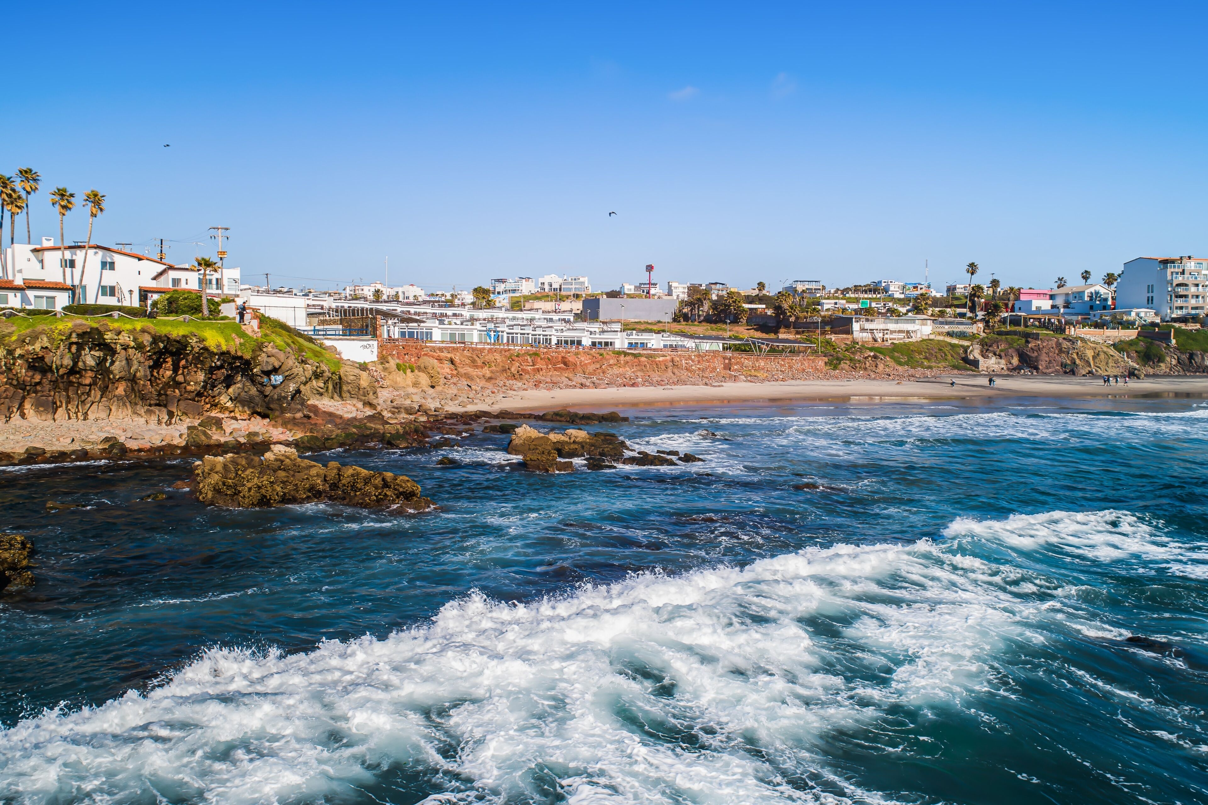 A coastal view with a rocky shoreline, waves crashing onto the beach, and buildings in the background.