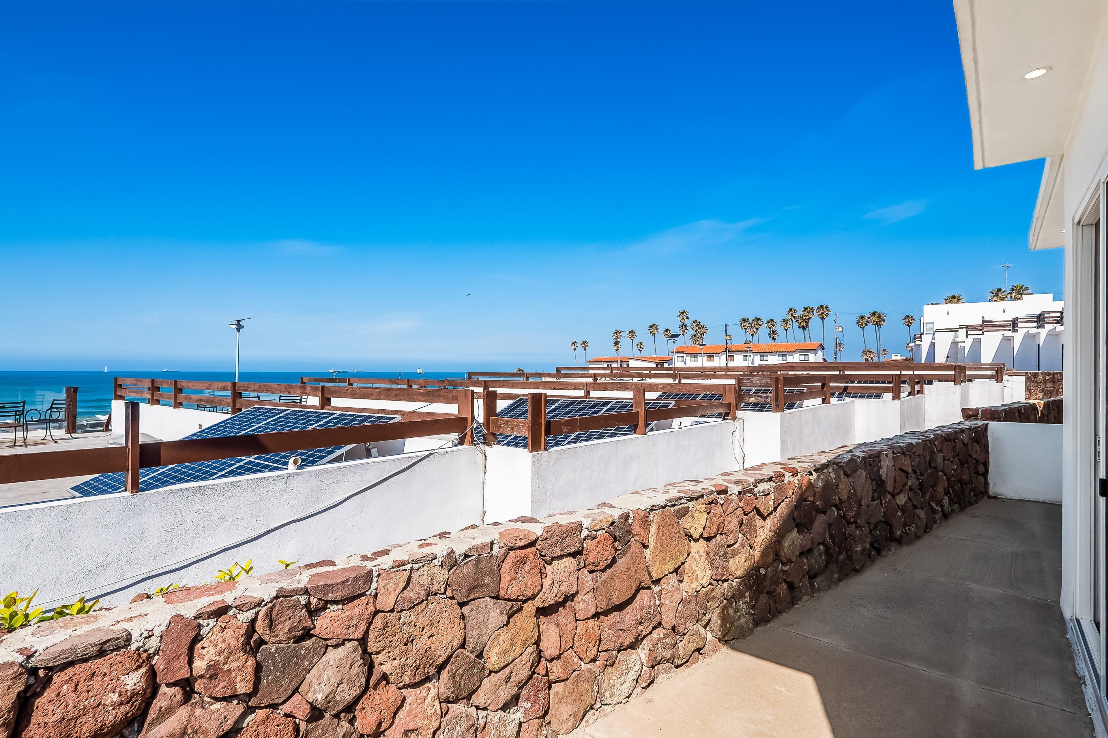 A rooftop terrace with solar panels, a stone wall, and a view of the ocean and palm trees.